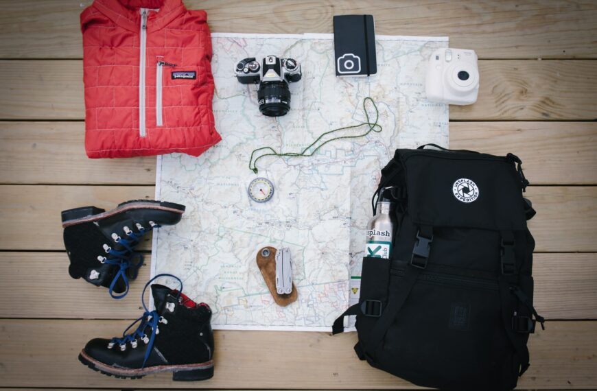 black hiking backpack near white Fujifilm instax mini camera near black leather boots, red half-zip jacket, gray pocket watch on white map